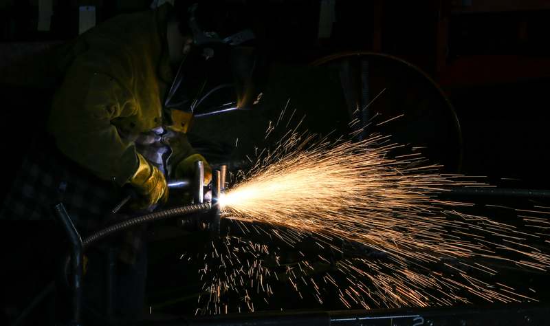 Close-up of a handheld plasma cutter in action on a steel plate, sparks flying, set in a well-lit metalworking workshop with clamps and safety gear visible